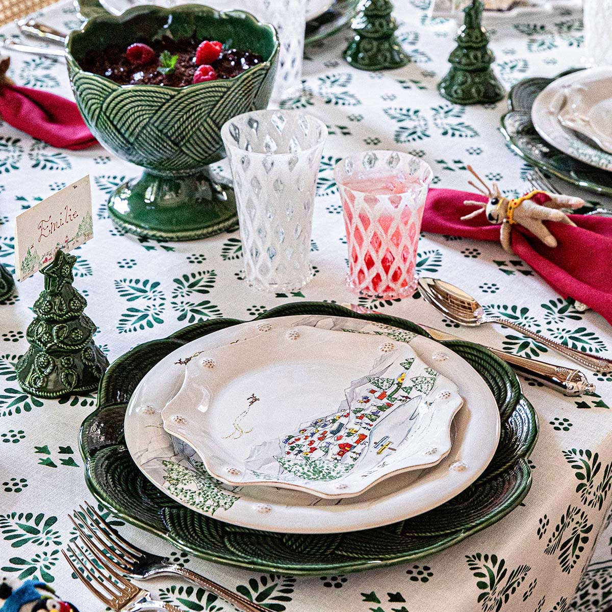 A festive table setting showcases Juliska's Berry & Thread North Pole Dinnerware and Serveware Collection, alongside holiday glassware, red napkins, small tree figurines, and a patterned tablecloth.