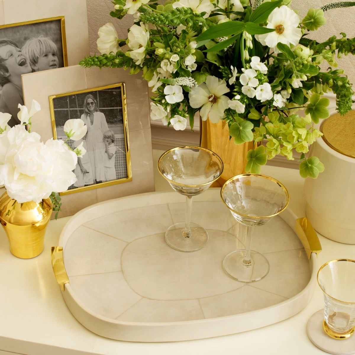 The Livio Parchment Serving Tray by Aerin, featuring gold handles, displays two empty martini glasses and is surrounded by gold vases with white flowers and framed black-and-white family photos.