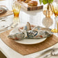 A neatly set dining table featuring the Kim Seybert Whistler Placemat, with a patterned napkin on a white plate, gold utensils, amber glasses, and croissants on a wooden board adding rustic charm.