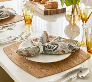 A neatly set dining table featuring the Kim Seybert Whistler Placemat, with a patterned napkin on a white plate, gold utensils, amber glasses, and croissants on a wooden board adding rustic charm.