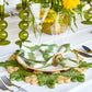A table setting with green and gold accents features Kim Seybert's Verve Placemat in Green & Natural, a patterned napkin with a gold ring, elegant glassware, and a floral centerpiece with yellow flowers.