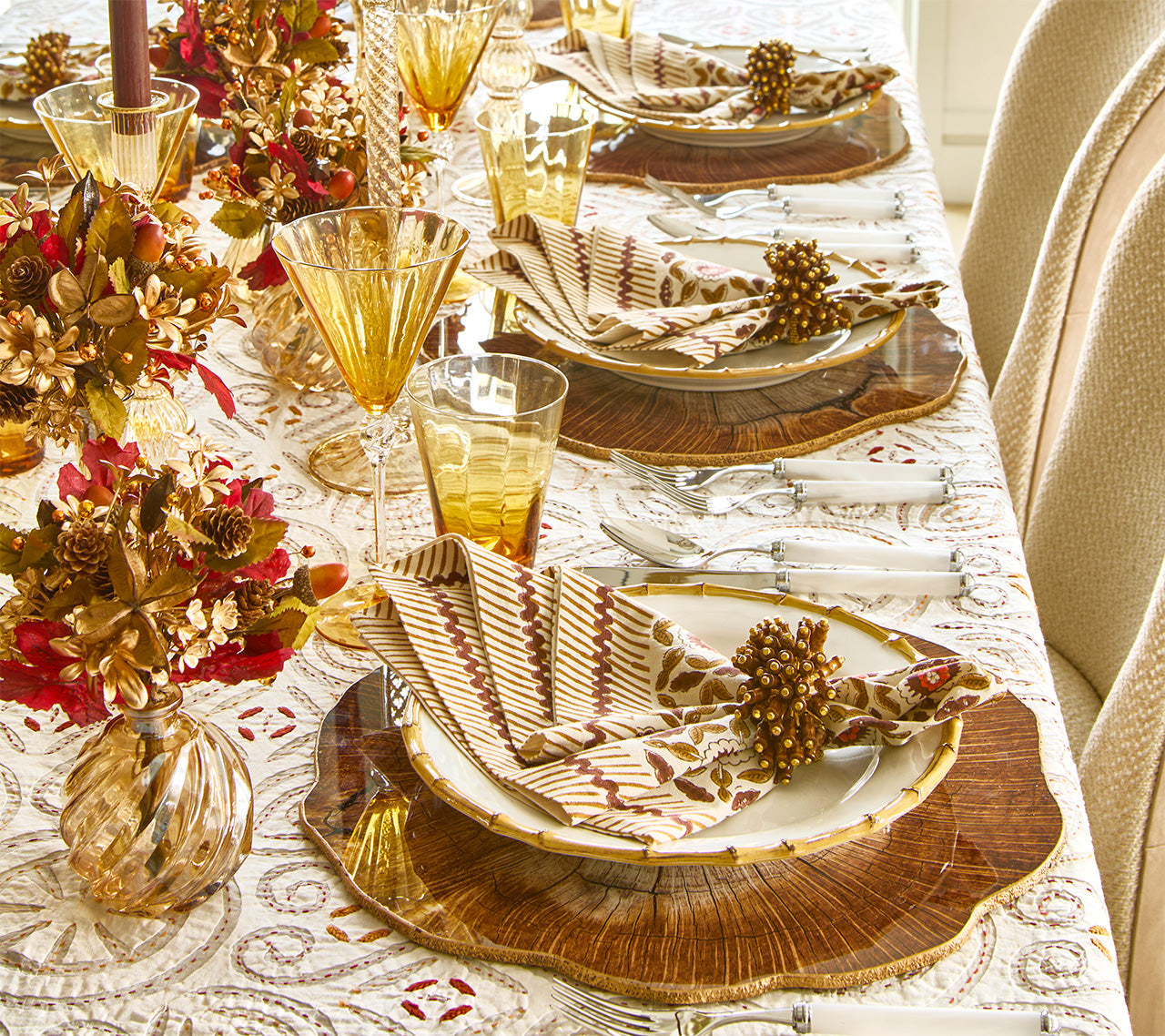 A formal dining table set with Kim Seybert Majorelle Napkin Set, gold-accented glasses, autumn-themed centerpieces, and layered plates on woven placemats.