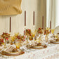 A formal dining table features gold glassware, brown candles, floral centerpieces, pinecones, and patterned plates on the elegant Malta Tablecloth Collection by Kim Seybert.
