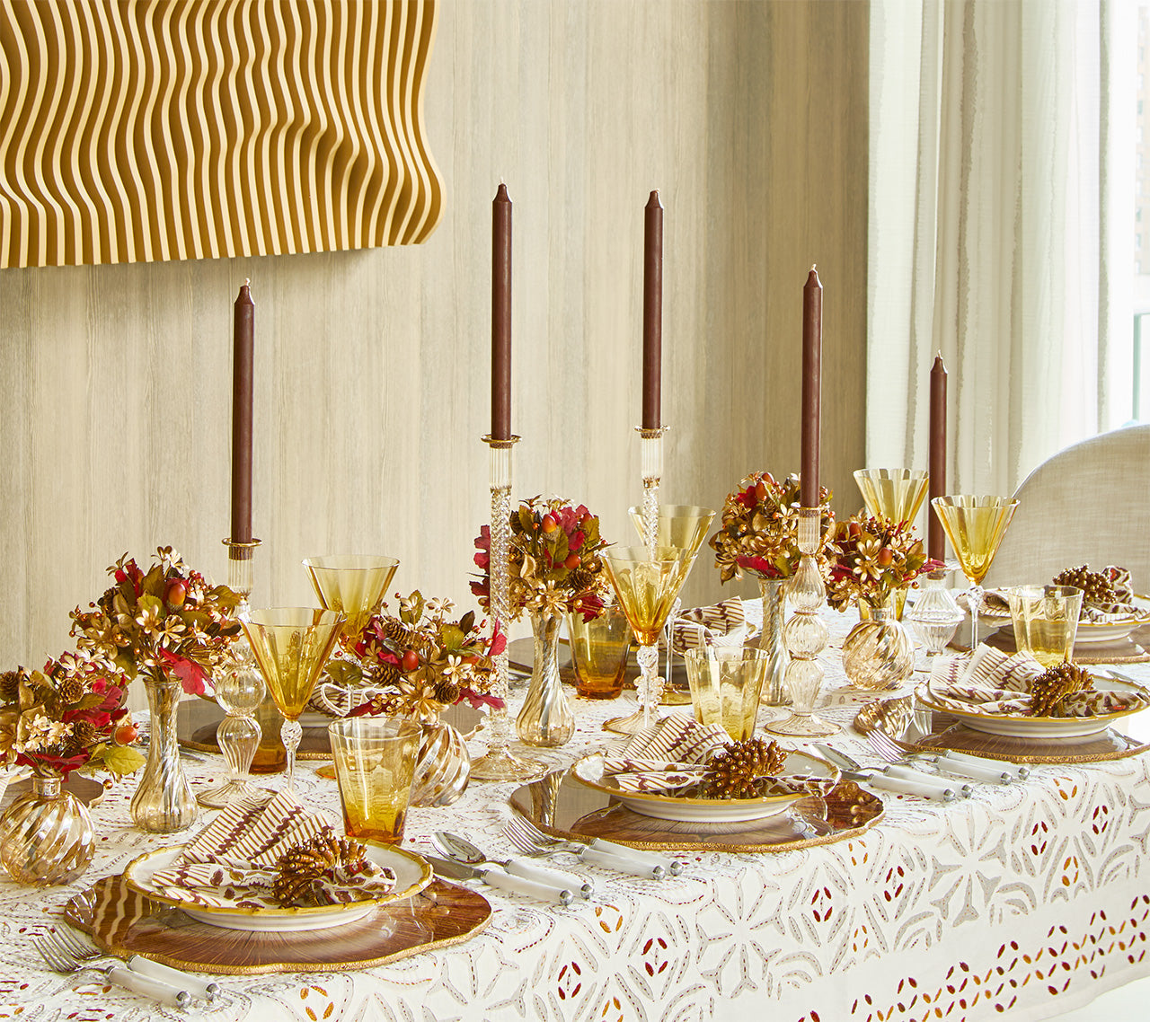 A formal dining table features gold glassware, brown candles, floral centerpieces, pinecones, and patterned plates on the elegant Malta Tablecloth Collection by Kim Seybert.