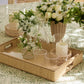 The Colette Cane Tray by Aerin holds two glasses, a carafe, a small vase with white flowers, and a candle; more flowers and another candle sit on a green and white patterned tablecloth.