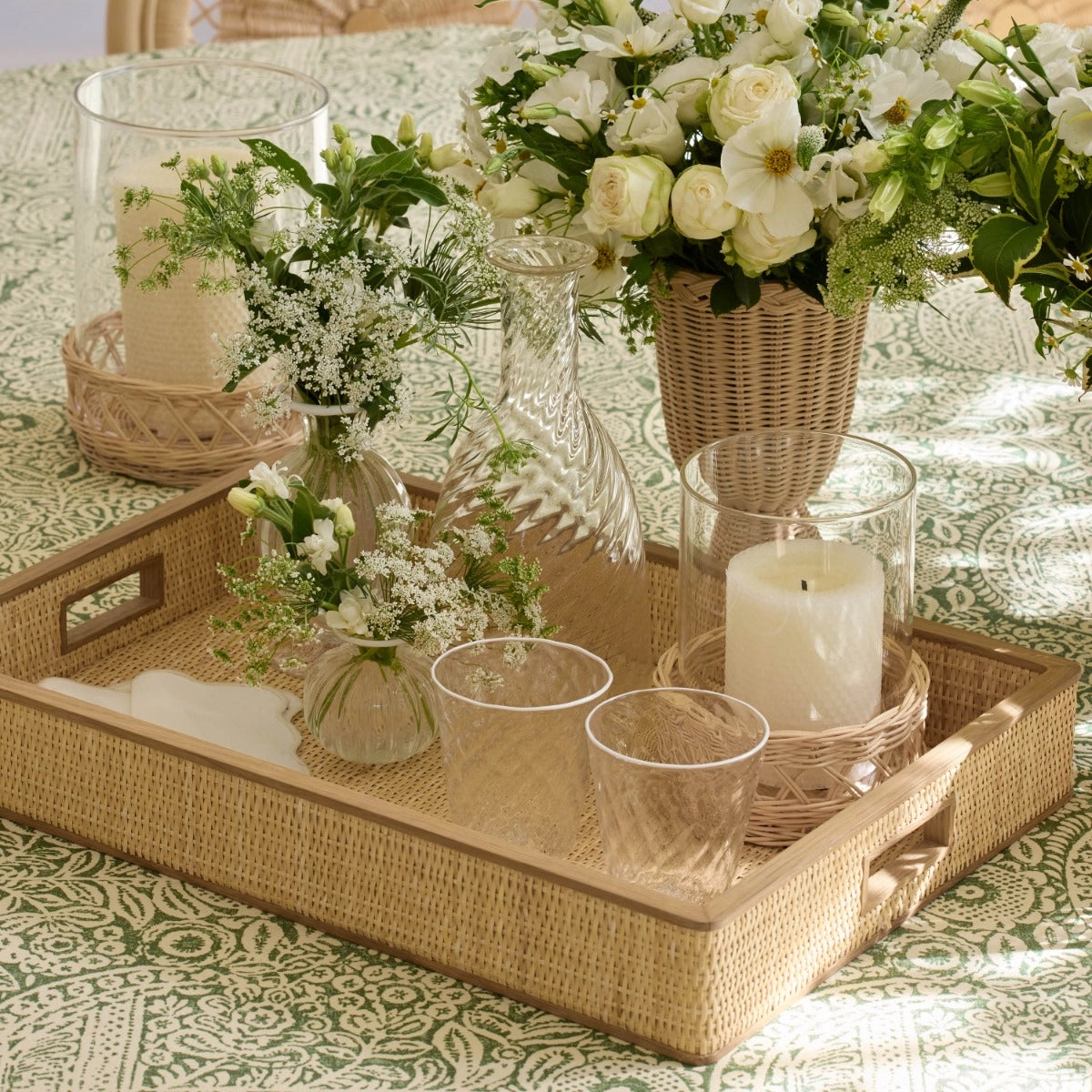 The Colette Cane Tray by Aerin holds two glasses, a carafe, a small vase with white flowers, and a candle; more flowers and another candle sit on a green and white patterned tablecloth.