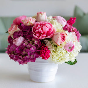 The Live Mother's Day Floral Arrangement by Hive Floral Studio features pink, purple, and white roses, hydrangeas, and peonies in a white ceramic vase, displayed on a white surface with a blurred background.