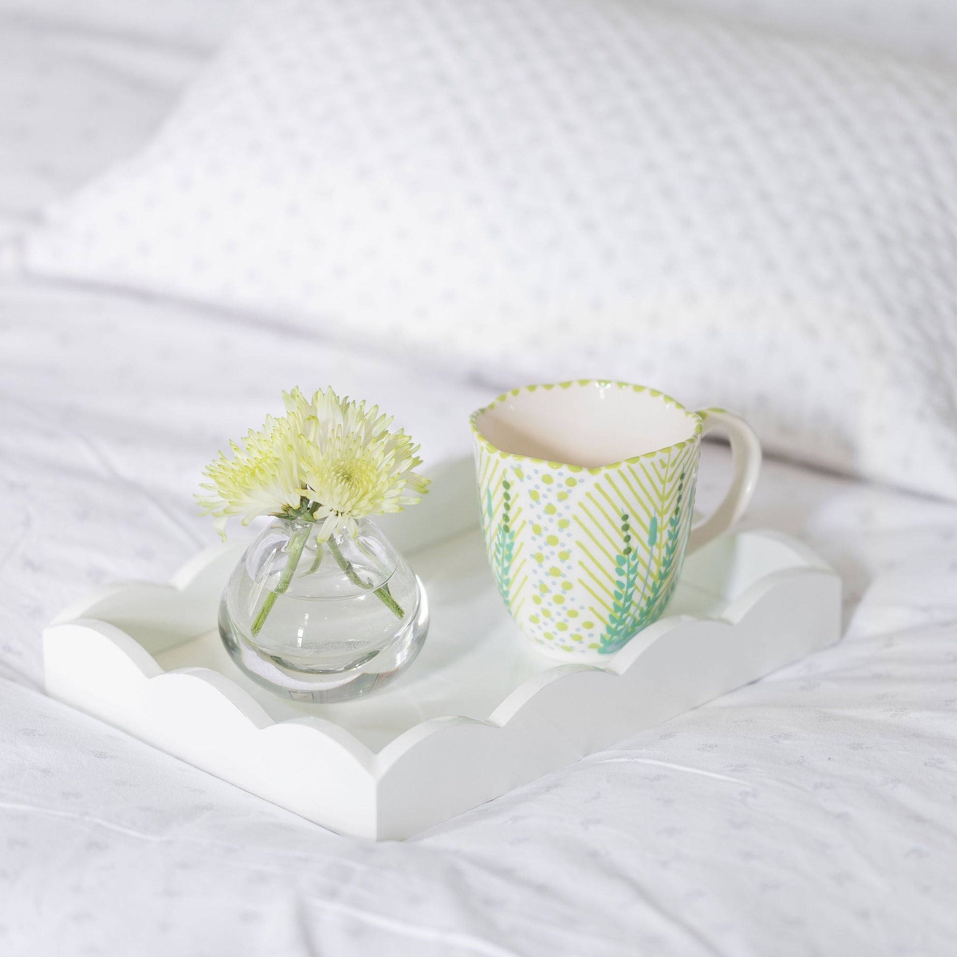 A scalloped white lacquer tray from Addison Ross holds a small glass vase with white flowers and a patterned mug atop a white bed with textured pillows.