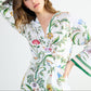 Yves Delorme x Laurence Tavernier Botanic Dressing Gown in white cotton sateen with a colorful botanical print, green-striped cuffs, and a wrap-style tie waist.