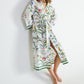 A person wearing a floor-length Yves Delorme x Laurence Tavernier Botanic Dressing Gown in a white cotton sateen floral print, patterned with colorful botanical illustrations and birds.