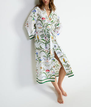 A person wearing a floor-length Yves Delorme x Laurence Tavernier Botanic Dressing Gown in a white cotton sateen floral print, patterned with colorful botanical illustrations and birds.