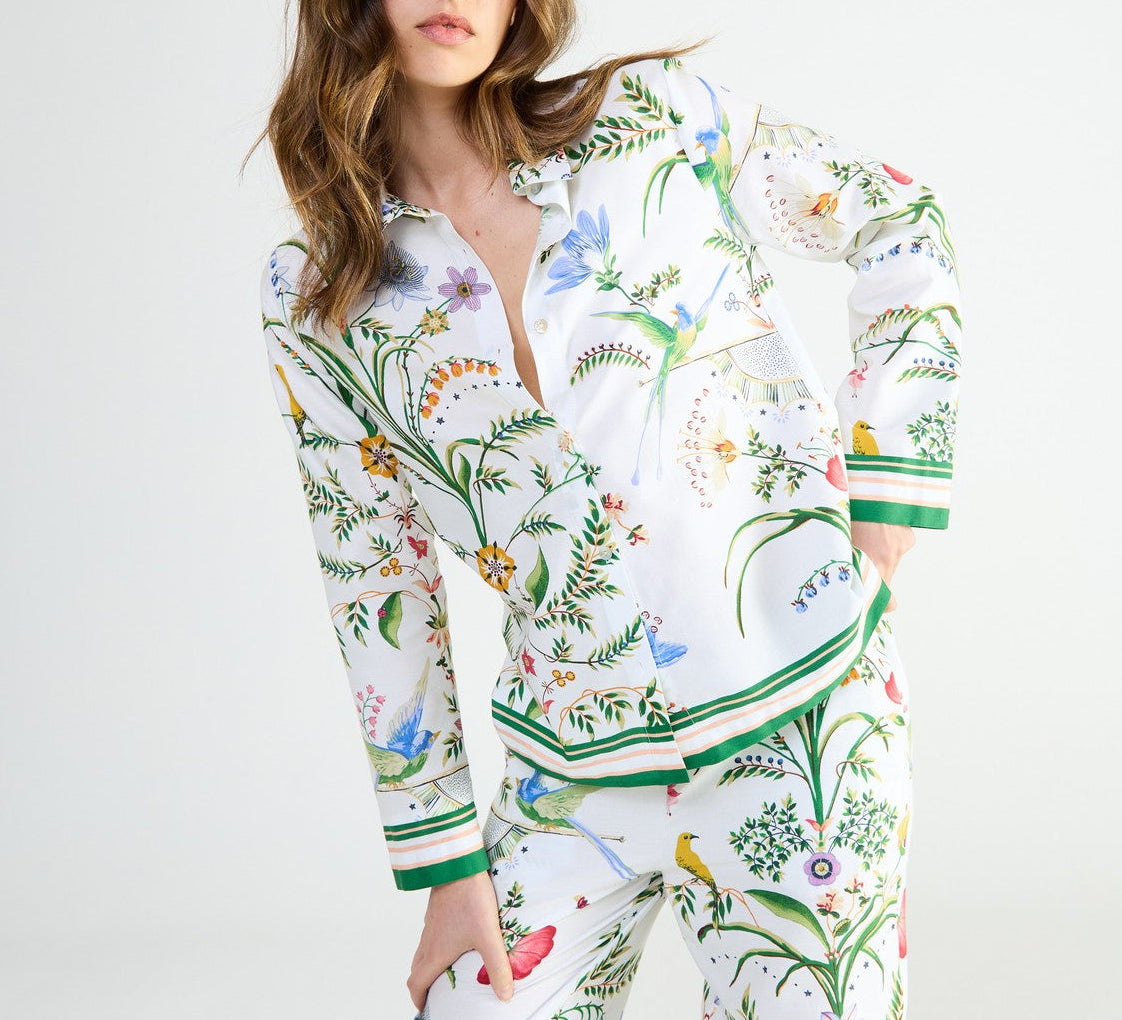 Yves Delorme x Laurence Tavernier Botanic pajamas consisting of a long-sleeved button-down top and matching pants in a floral print with colorful flowers, birds, and insects on a white background.