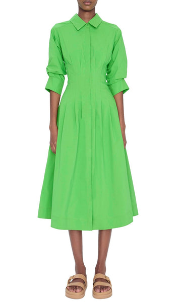A woman wearing a bright green, midi-length Simkhai Signature Jazz dress with short sleeves and a button-up front closure, paired with light brown sandals.
