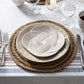 A table setting featuring the Juliska Le Panier Placemat, stacked beige and white plates, silverware, glassware, a cup, and a decorative knotted rope accent arranged atop the plates.