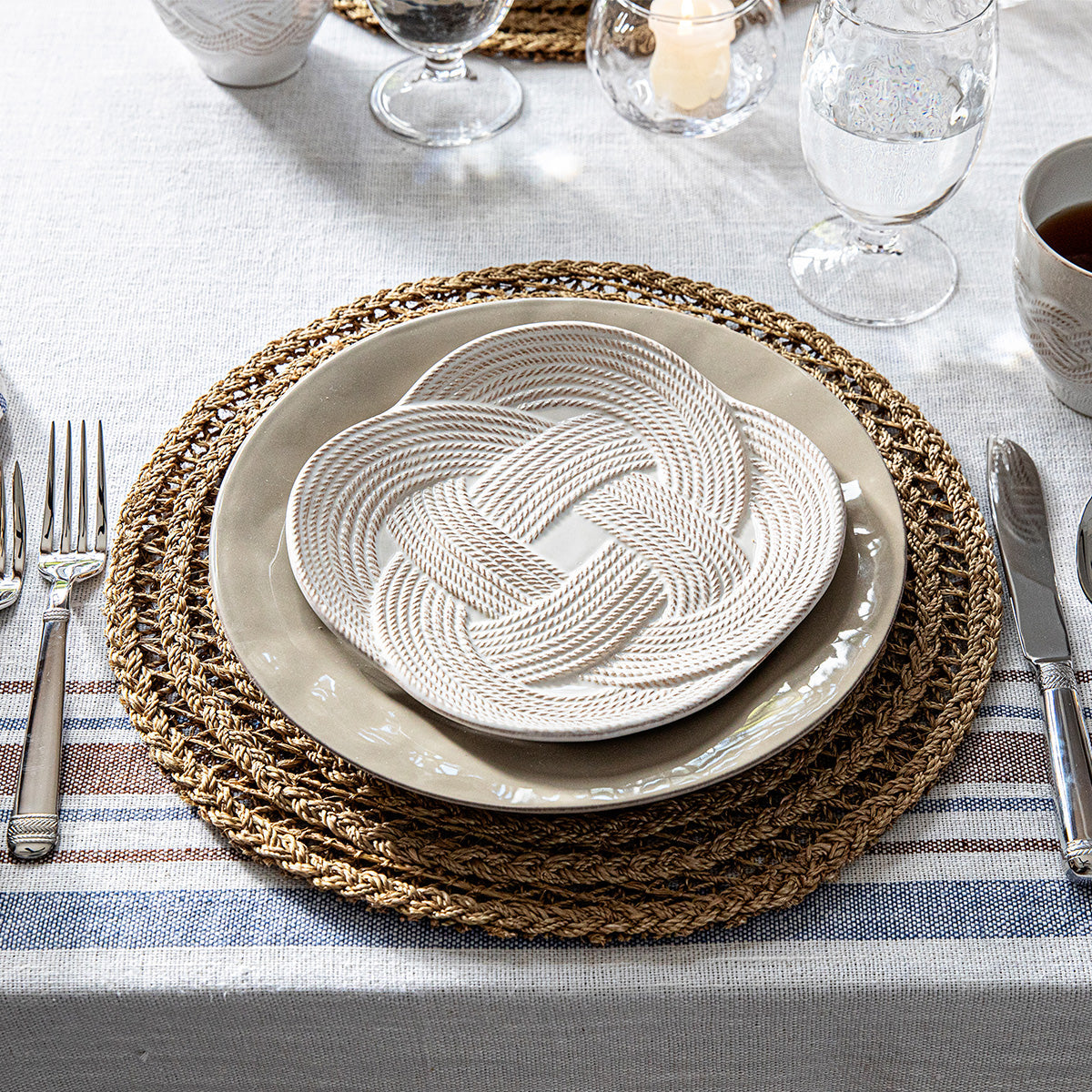 A table setting featuring the Juliska Le Panier Placemat, stacked beige and white plates, silverware, glassware, a cup, and a decorative knotted rope accent arranged atop the plates.