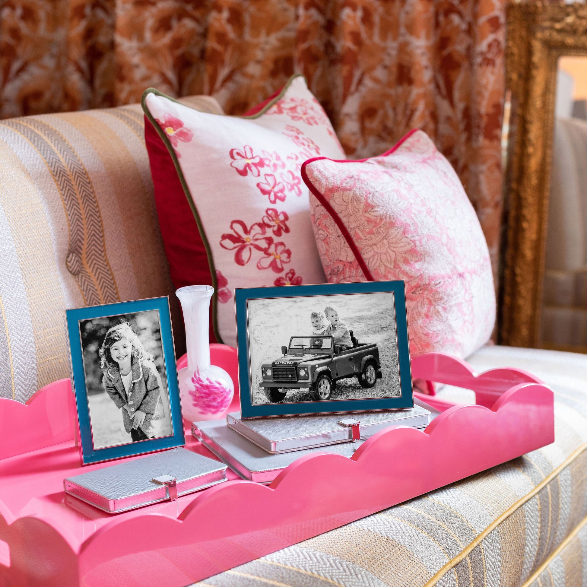 On a striped sofa, the Addison Ross Scalloped Watermelon Pink Lacquer Tray holds two framed photos, a small glass vase, and two books; patterned pillows and a curtain appear in the background.