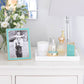 A white dresser displays an Addison Ross Gold Trim Enamel Frame, a tray of perfumes, a candle, and a lamp, with a vase of white flowers in the background.
