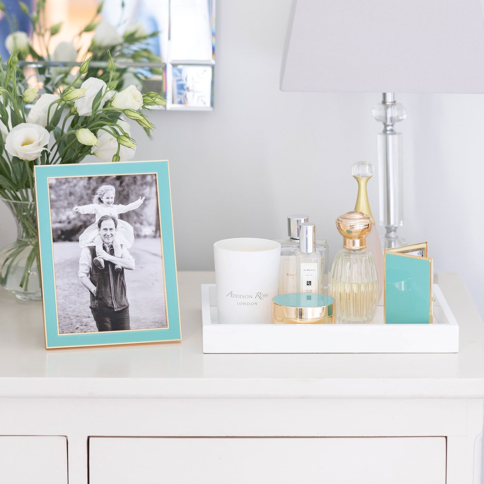 A white dresser displays an Addison Ross Gold Trim Enamel Frame, a tray of perfumes, a candle, and a lamp, with a vase of white flowers in the background.