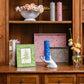 A wooden shelf displays books, candles, framed art, florals, and a blue candle in a white hand-shaped holder atop an Addison Ross Small Chinoiserie Tray Collection piece.