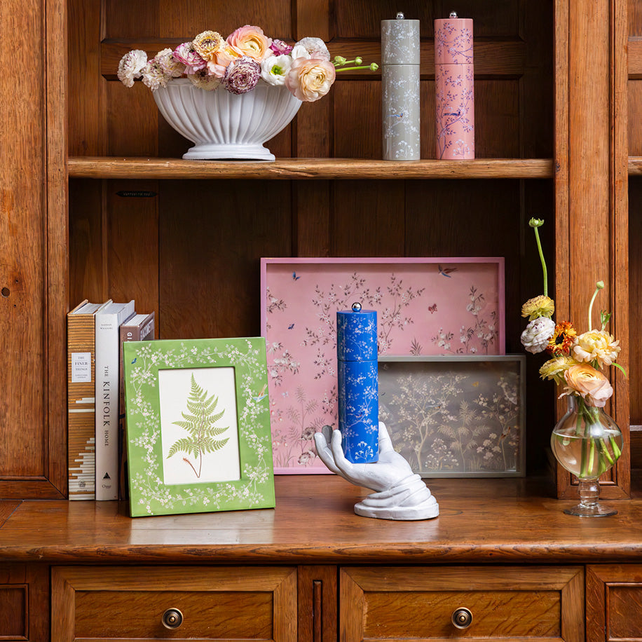 A wooden shelf displays books, candles, framed art, florals, and a blue candle in a white hand-shaped holder atop an Addison Ross Small Chinoiserie Tray Collection piece.