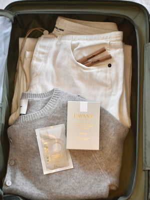 A packed suitcase with white pants, a white top, a gray sweater, two L'AVANT plant-based skincare products, two lip gloss tubes, and an L'Avant Collective Fresh Linen Laundry Detergent Travel Pack ensures you're adventure-ready and cruelty-free.