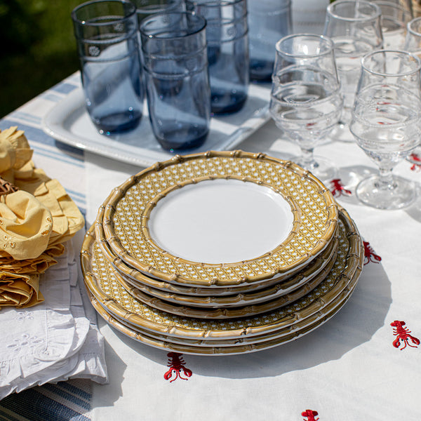 Juliska’s Bamboo Caning Melamine Dinnerware Collection is stacked on a white tablecloth with red embroidery, paired with yellow napkins, clear wine glasses, and blue tumblers for a vibrant tablescape.