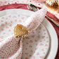A close-up of a table setting featuring Juliska’s Berry & Thread Heart Napkin Ring and Place Card Holder Collection—white plate with pink hearts, matching napkin, and hammered gold heart napkin ring.