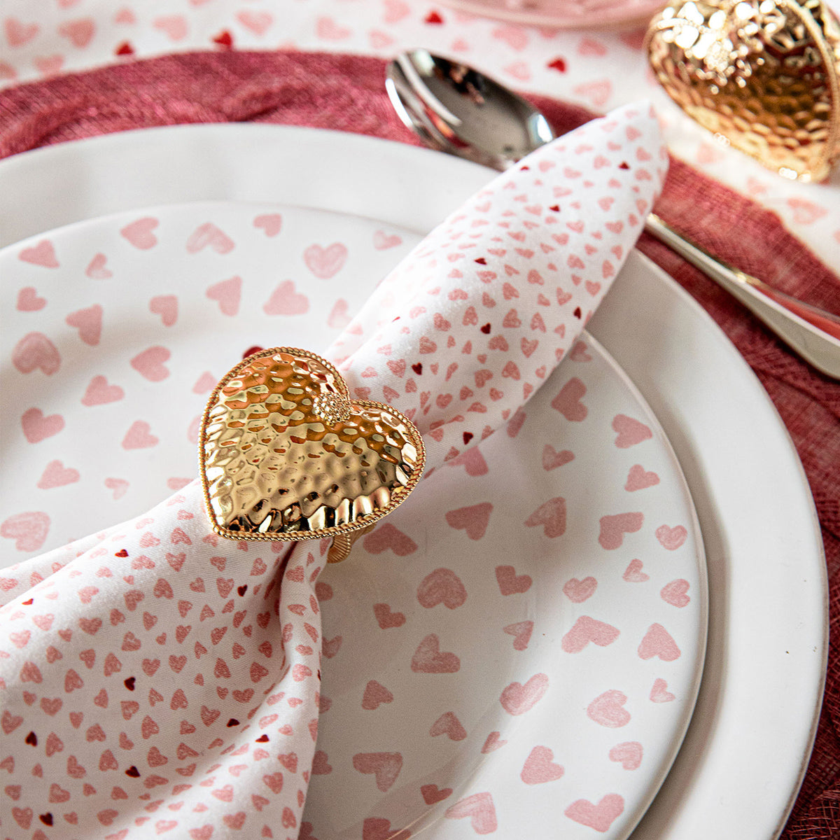 A close-up of a table setting featuring Juliska’s Berry & Thread Heart Napkin Ring and Place Card Holder Collection—white plate with pink hearts, matching napkin, and hammered gold heart napkin ring.