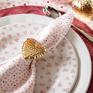 A close-up of a table setting featuring Juliska’s Berry & Thread Heart Napkin Ring and Place Card Holder Collection—white plate with pink hearts, matching napkin, and hammered gold heart napkin ring.