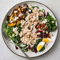 Enjoy a vibrant salad with shredded chicken, sliced mushrooms, peas, leafy greens, cherry tomatoes, wholesome grains, a quartered boiled egg, and a drizzle of dressing served on a white plate with a fork—an ideal dish inspired by *The Complete High-Protein Cookbook* from Penguin Random House for anyone looking to add high-protein meals to their balanced meal plans.