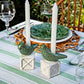 The Berry & Thread Bird Topiary Candlestick Set by Juliska features two white candles in green songbird-shaped holders, displayed on a striped tablecloth amidst glassware and green plates for a fresh spring tablescape.
