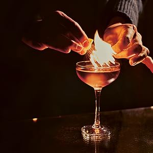 A person holds a match over a martini served in a coupe glass—referencing techniques celebrated by Chronicle Books in “The Martini”—igniting a flame above the drink in an intimate, low-lit setting.