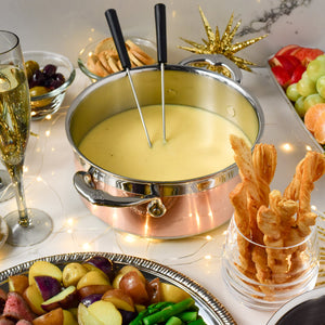A Hestan CopperBond Copper Induction Soup Pot, 3qt, filled with cheese fondue, is surrounded by breadsticks, vegetables, fruit, crackers, olives, and champagne on a decorated table. Two dipping forks rest in the oven-safe pot, ready to share.
