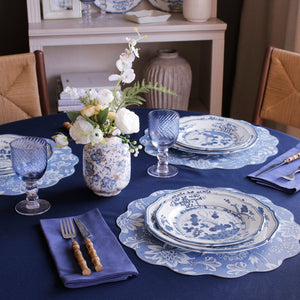 A round table features Beatriz Ball's Vida Faenza Melamine Dinnerware and Placemat Collection, blue glasses, bamboo-handled cutlery, navy napkins, and a vase of white flowers atop a blue tablecloth.