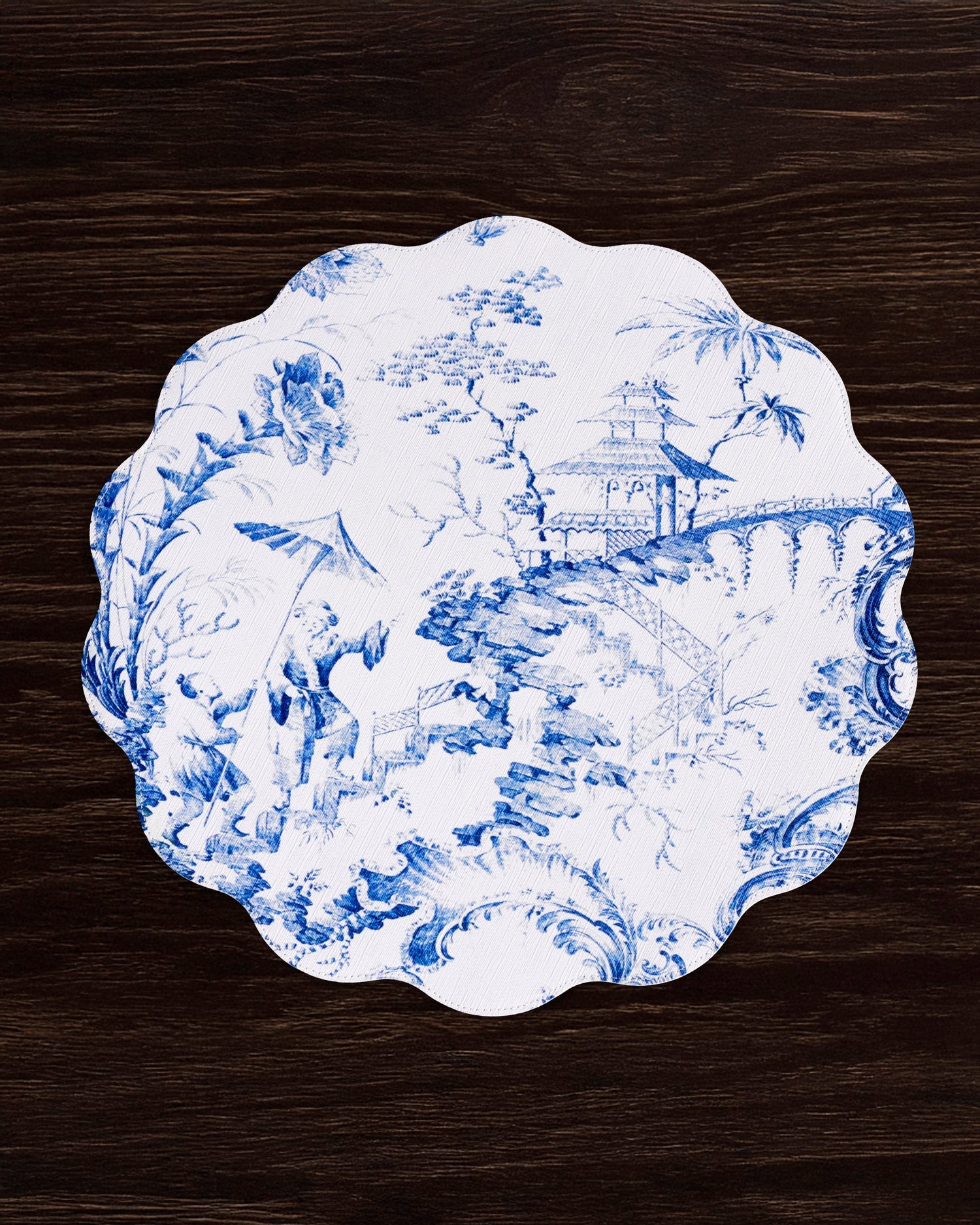 A Beatriz Ball VIDA Chinoiserie Reversible Round Placemat in blue and white holds a white plate with blue oriental-style designs, beautifully displayed on a dark wooden surface.