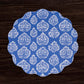 The Beatriz Ball VIDA Chinoiserie Reversible Round Placemat, featuring blue and white floral patterns with scalloped edges, is displayed on a dark wood surface.
