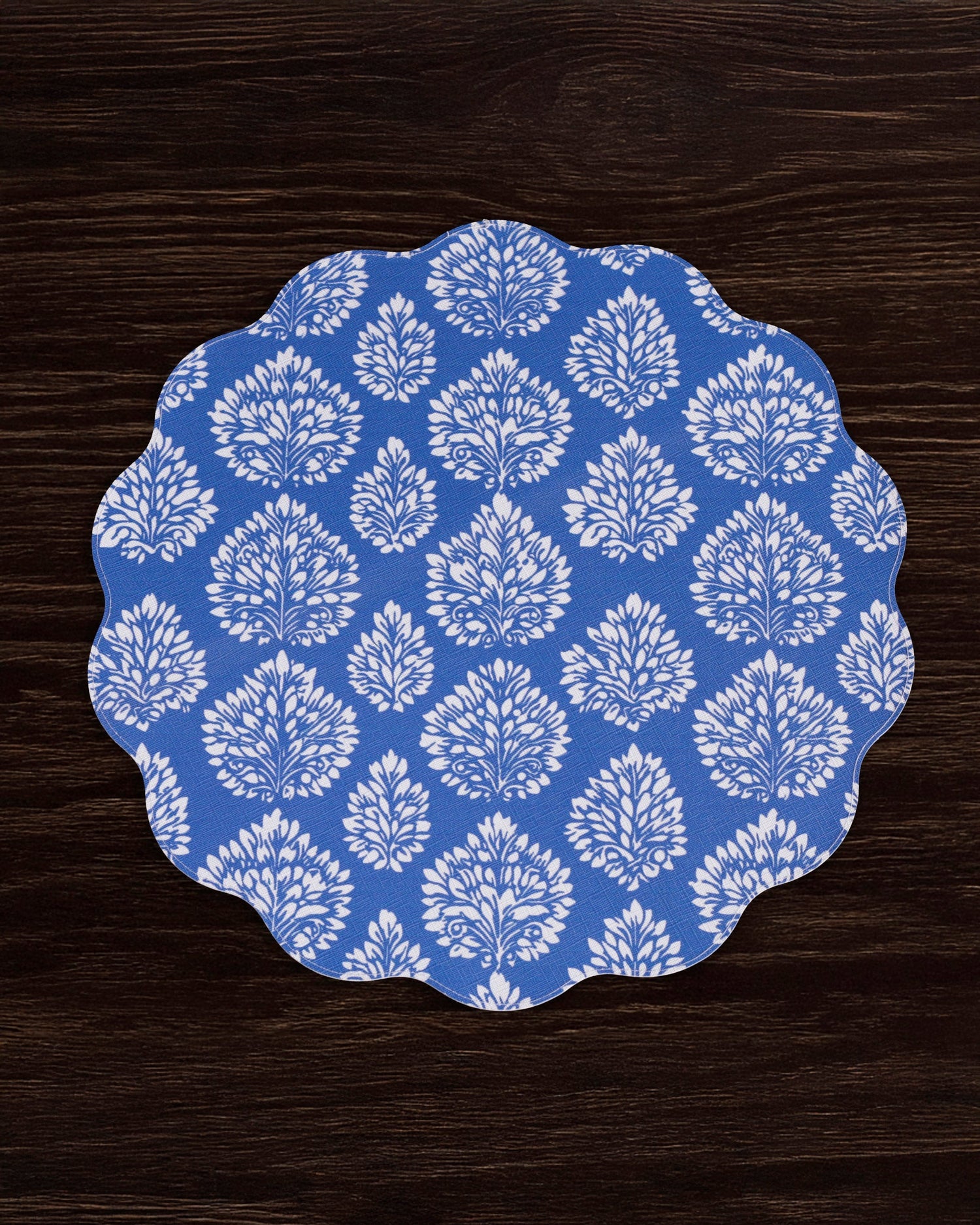 The Beatriz Ball VIDA Chinoiserie Reversible Round Placemat, featuring blue and white floral patterns with scalloped edges, is displayed on a dark wood surface.