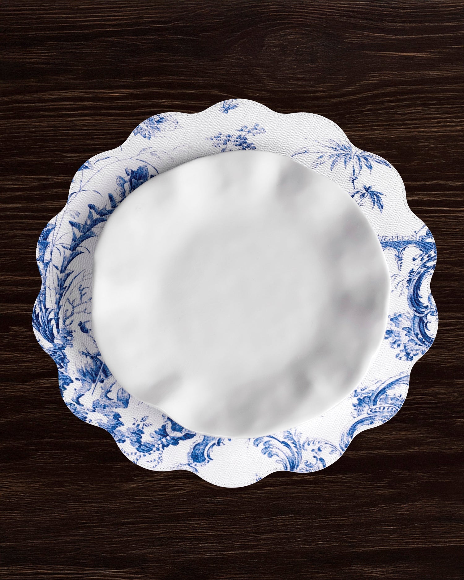 A white porcelain plate with a blue floral design rests on a matching scalloped charger, accented by Beatriz Ball’s VIDA Chinoiserie Reversible Round Placemats in blue and white on a dark wood table.
