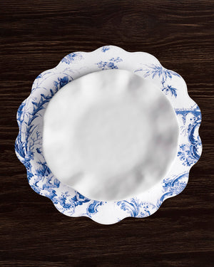 A white porcelain plate with a blue floral design rests on a matching scalloped charger, accented by Beatriz Ball’s VIDA Chinoiserie Reversible Round Placemats in blue and white on a dark wood table.