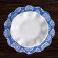 A round, white plate with a blue and white patterned, scalloped edge rests on a dark wooden surface, beautifully complemented by the Beatriz Ball VIDA Chinoiserie Reversible Round Placemat.