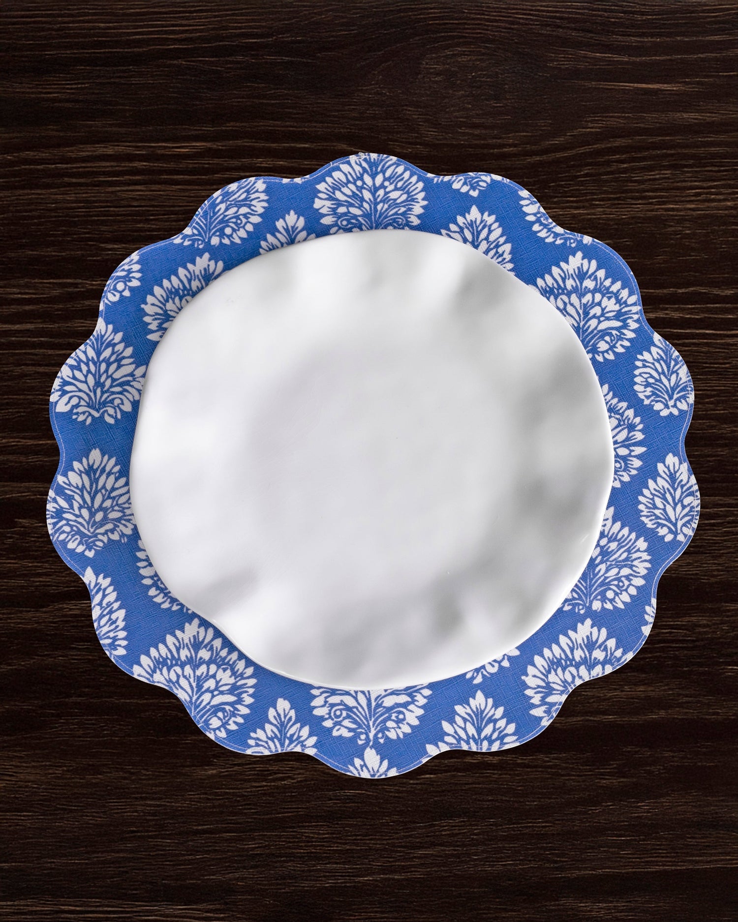 A round, white plate with a blue and white patterned, scalloped edge rests on a dark wooden surface, beautifully complemented by the Beatriz Ball VIDA Chinoiserie Reversible Round Placemat.