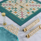 The WS Game Company's Scrabble Del Mar Shagreen Edition features a game board where letter tiles form words. A set of letter tiles, a bag, a sand timer, and two racks with tiles spelling "PLAY ME" and "WINNER" elegantly rest on a faux-shagreen marble surface, adding luxurious textures and premium components to the scene.