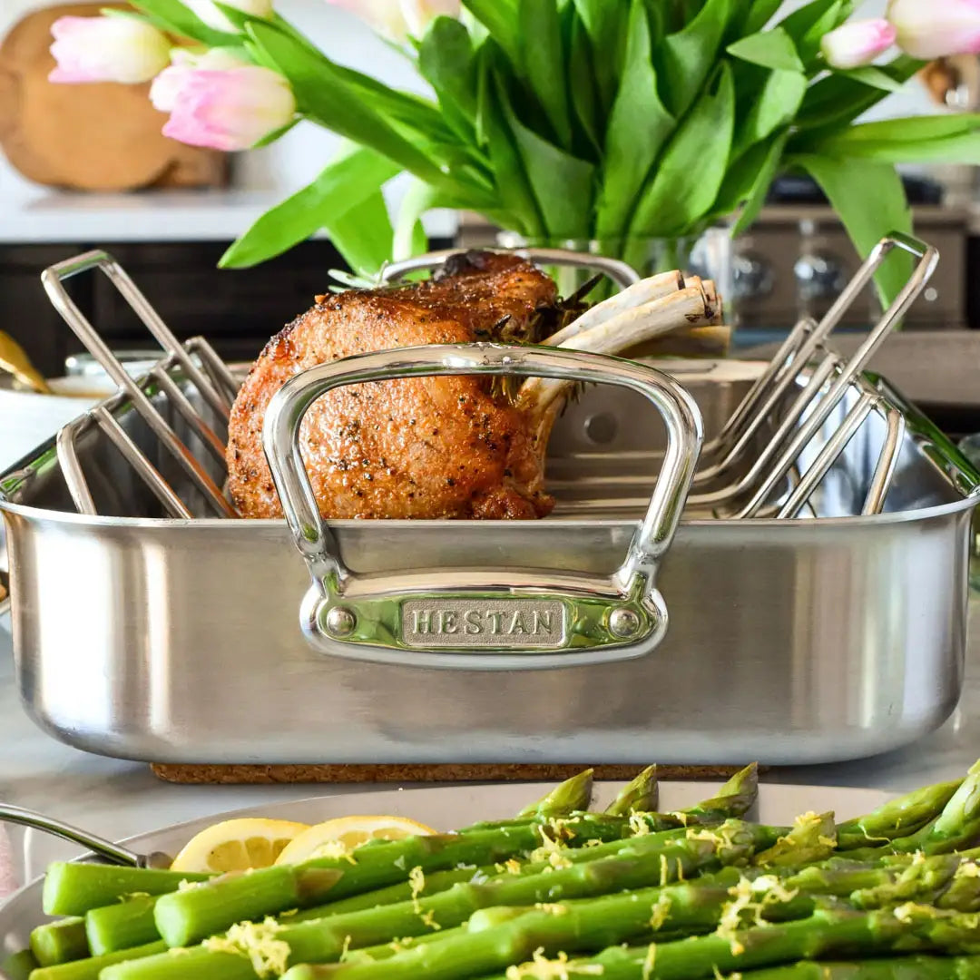 A roasted rack of meat in the Hestan Provisions Classic Clad Roaster with Rack Collection, paired with asparagus and lemon in the foreground, flowers visible in the background.