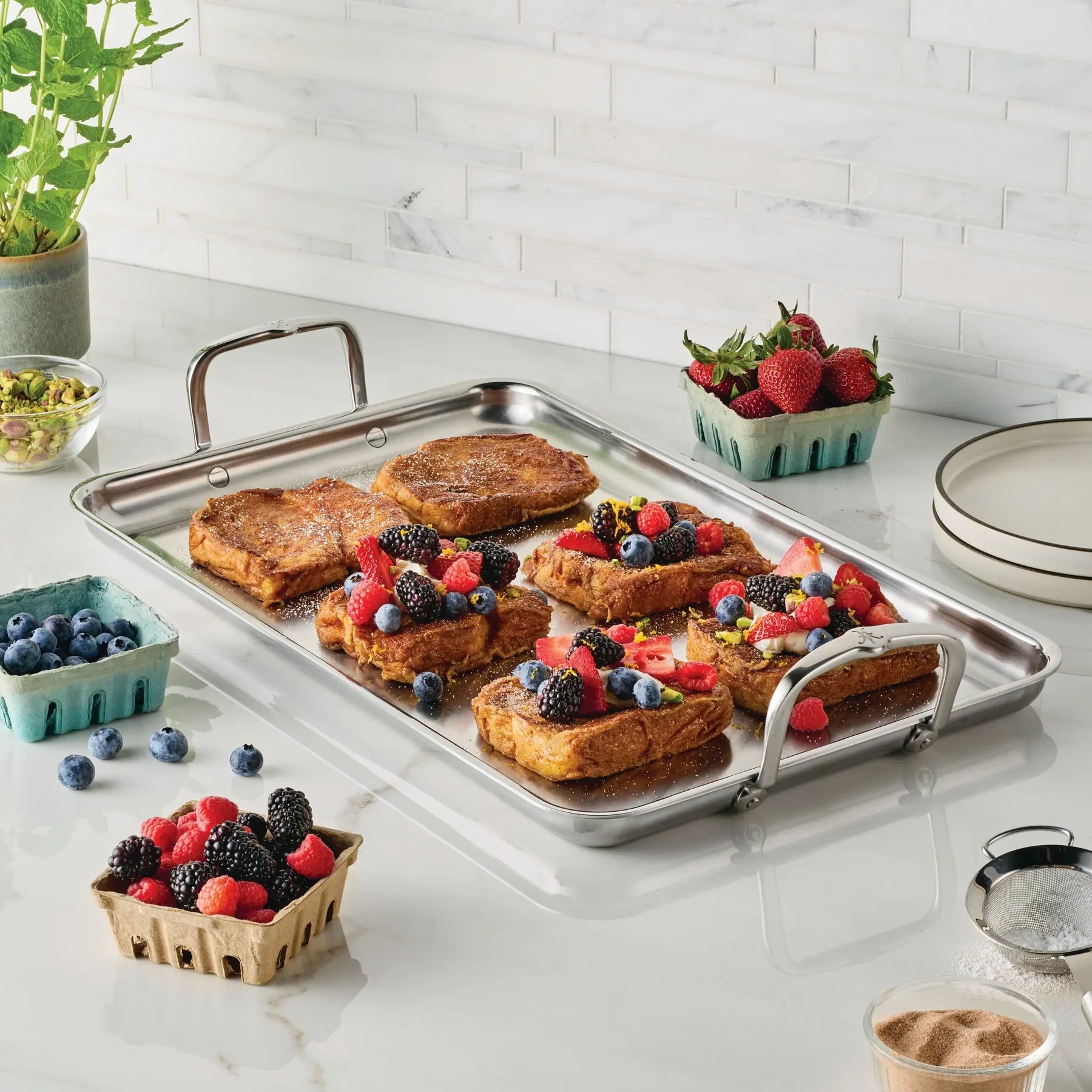 A stainless steel tray of French toast with berries and citrus zest rests on a kitchen counter beside the Hestan Provisions Double Burner Griddle, known for even heat distribution, with assorted berries and a small plant nearby.