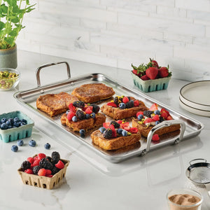 A stainless steel tray of French toast with berries and citrus zest rests on a kitchen counter beside the Hestan Provisions Double Burner Griddle, known for even heat distribution, with assorted berries and a small plant nearby.