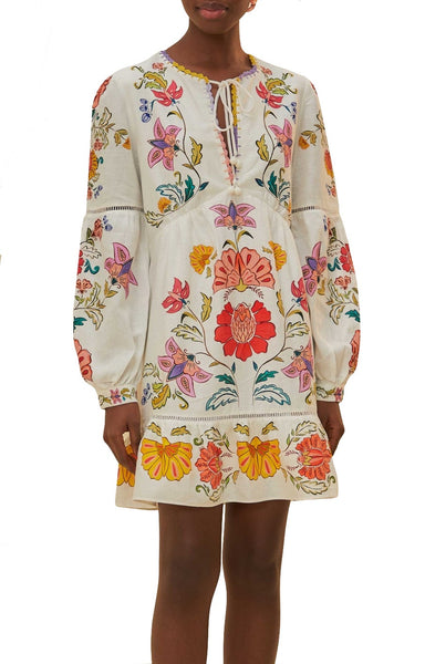 A woman wearing a Farm Rio Floral Insects off-white mini dress with long sleeves and a tie detail at the neckline, featuring botanical and insect embroidery.