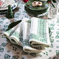 A set of Juliska’s Iberian Evergreen Border Napkins, neatly folded atop a stack of green plates, adds an elegant touch to the meal setting on a patterned tablecloth.