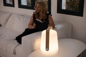 A woman reads on a white sofa while the Fermob Bamboo Balad 15" Lamp—ideal for portable lighting and outdoor use—glows on a nearby white table in the foreground.