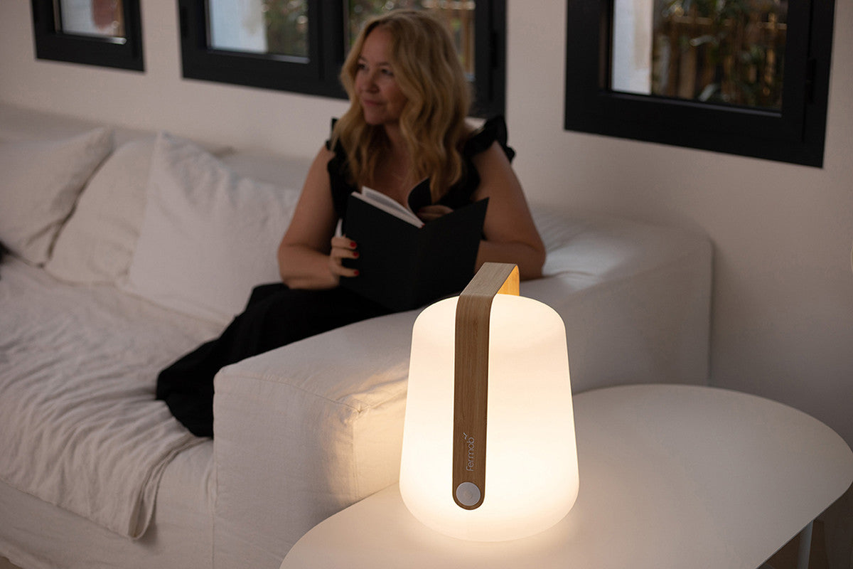 A woman sits on a white sofa reading, while a Fermob Bamboo Balad 10" Lamp with a wooden handle casts a warm, portable glow on the nearby table.