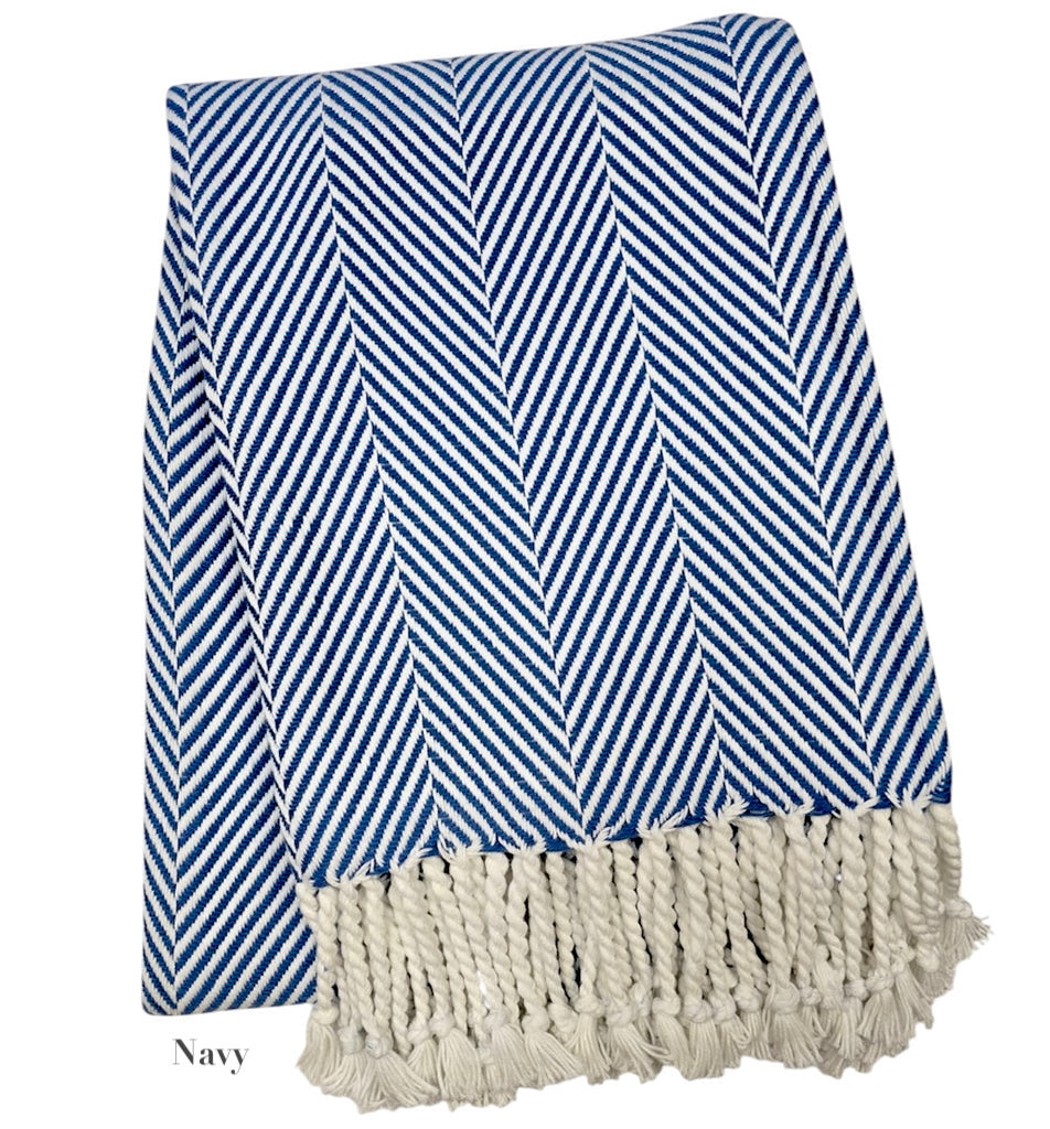 The Nantucket Herringbone Throw Collection by A Soft Idea features a folded navy and white throw with hand-knotted fringe, classic herringbone pattern, and soft cotton craftsmanship.